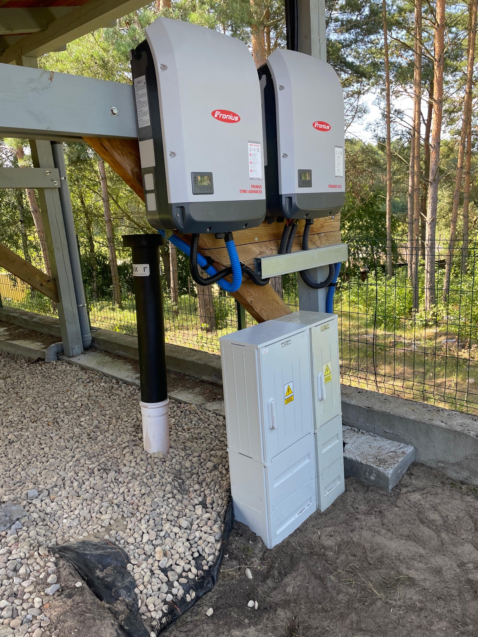 Dwa inwertery Fronius zamontowane na drewnianej konstrukcji obok białej skrzynki elektrycznej na tle lasu. Widoczne niebieskie kable i kamieniste podłoże.