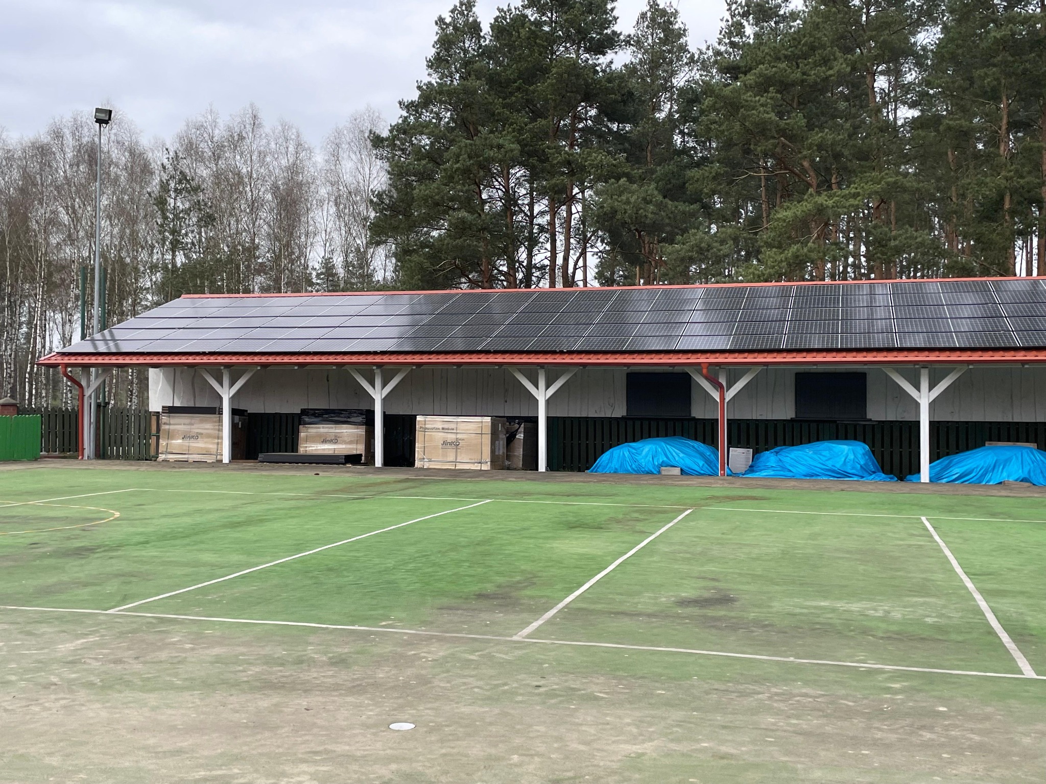 Zadaszenie boiska sportowego z panelami fotowoltaicznymi na dachu, widoczne skrzynie z panelami Jinko i niebieskie plandeki. Tło stanowią drzewa.