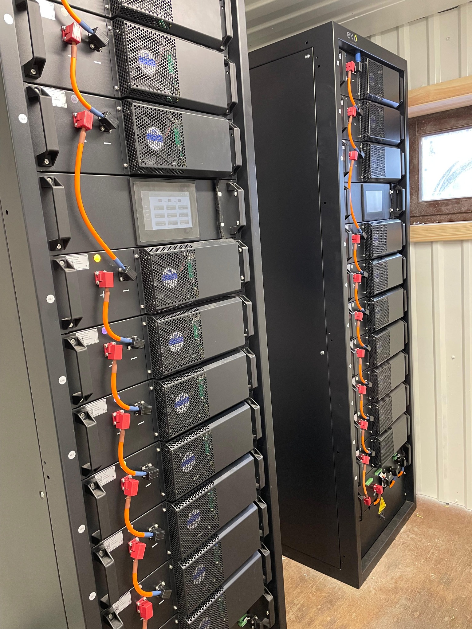Dwa czarne regały z modułami magazynu energii EKT, połączone pomarańczowymi kablami, widoczne wentylatory i ekrany dotykowe, w tle okno w pomieszczeniu.
