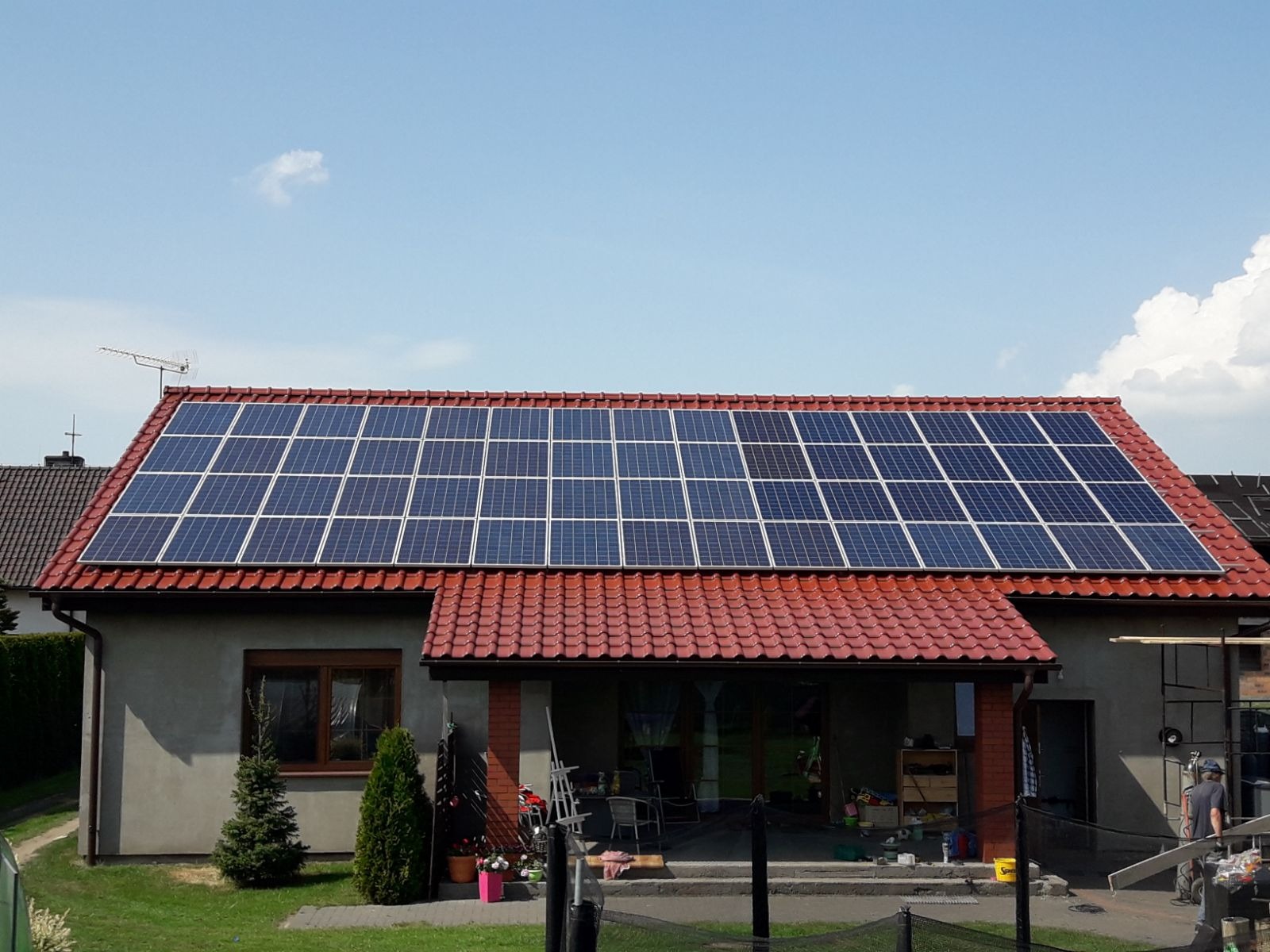Dom jednorodzinny z panelami fotowoltaicznymi na czerwonym dachu, widok z ogrodu w słoneczny dzień. Antena telewizyjna na szczycie dachu.
