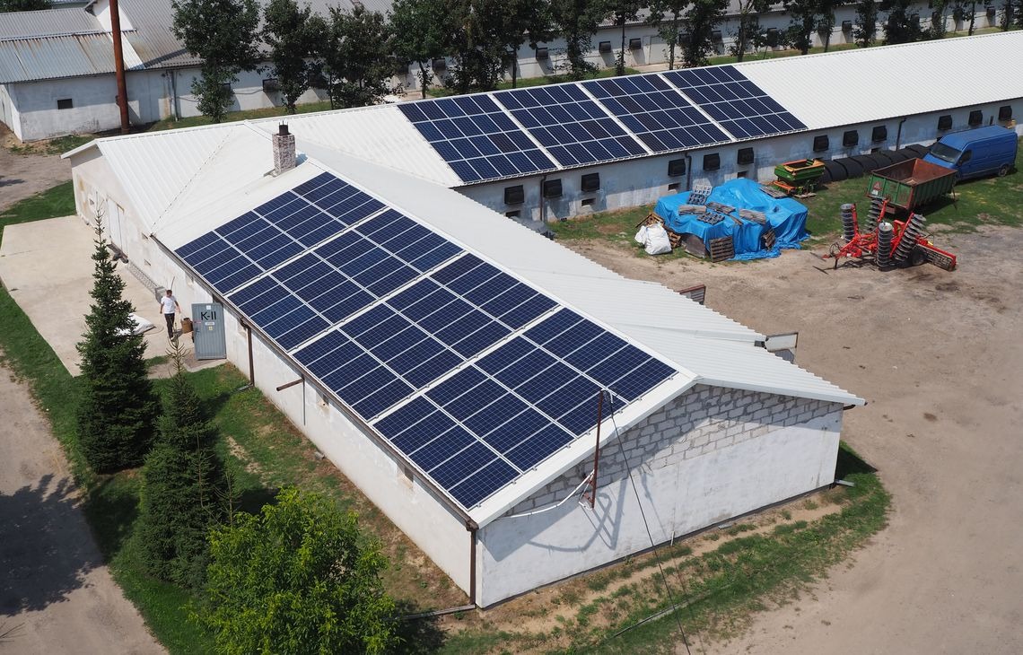 Widok z góry na budynek gospodarczy z panelami fotowoltaicznymi na dachu. Obok zaparkowane maszyny rolnicze i osoba wchodząca do budynku.
