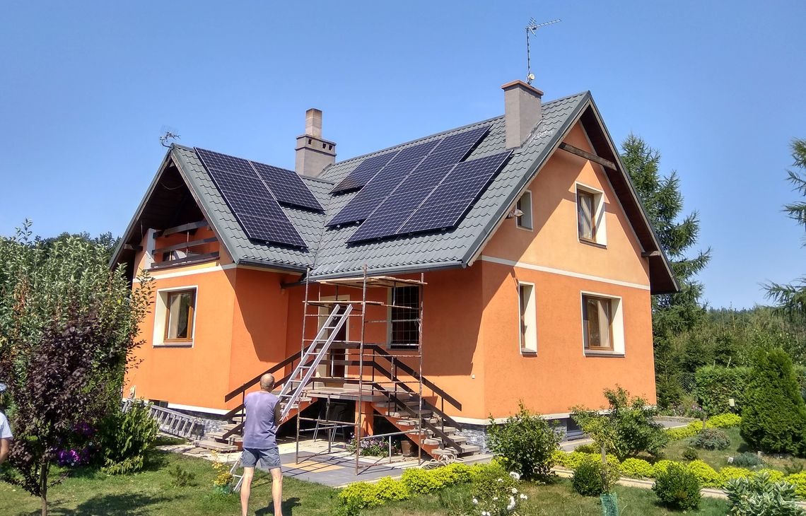 Pomarańczowy dom jednorodzinny z szarym dachem, na którym zamontowano panele fotowoltaiczne. Na pierwszym planie rusztowanie i osoba pracująca przy instalacji.