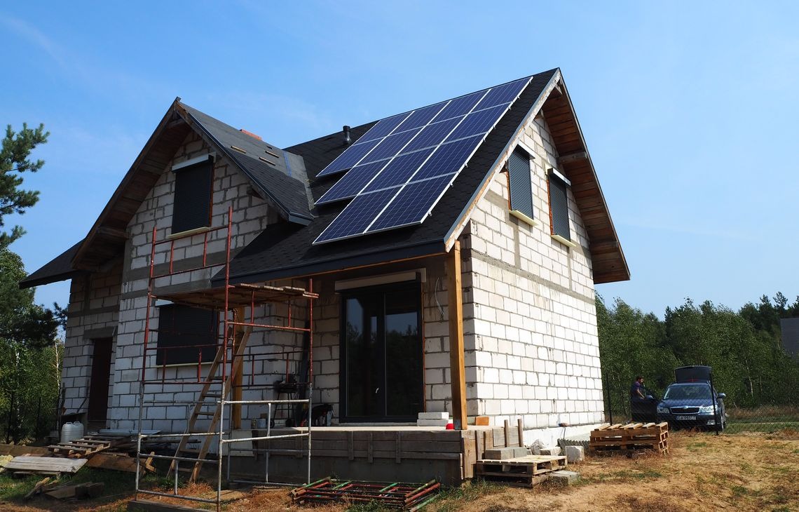 Dom w trakcie budowy z zamontowanymi panelami fotowoltaicznymi na dachu. Widoczne rusztowania i materiały budowlane. Samochód osobowy z otwartym bagażnikiem w tle.