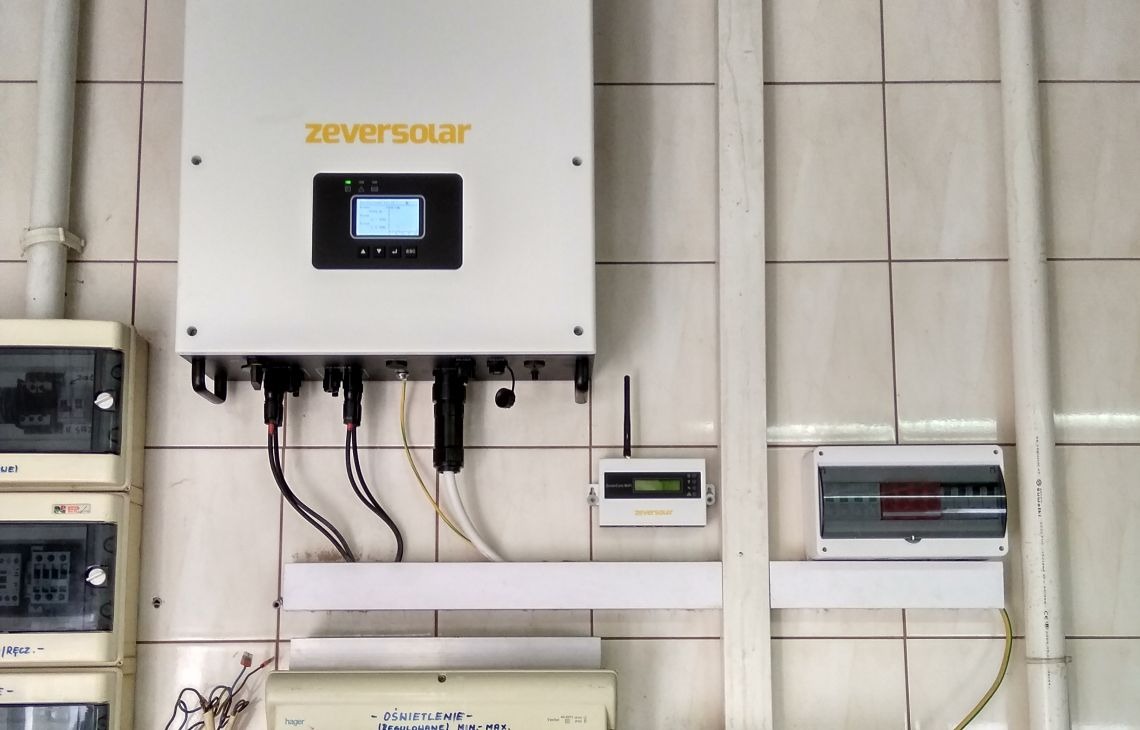 Biały inwerter Zeversolar zamontowany na kafelkowanej ścianie, widoczne przewody, skrzynki elektryczne i rury instalacyjne. Układ poziomy, oświetlenie dzienne.