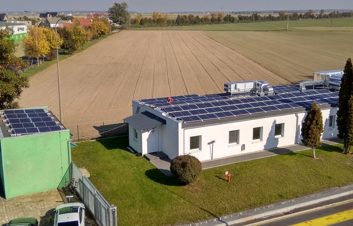 Instalacja fotowoltaiczna na dachu budynku z białymi ścianami, widok z góry na tle pola uprawnego i zabudowań. Osoba w czerwonej koszulce na dachu.