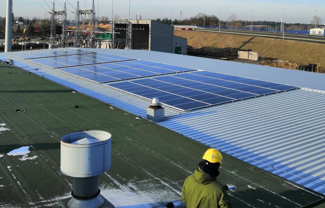 Instalacja paneli fotowoltaicznych na dachu przemysłowym z widoczną osobą w kasku, w tle słupy wysokiego napięcia i droga ekspresowa.