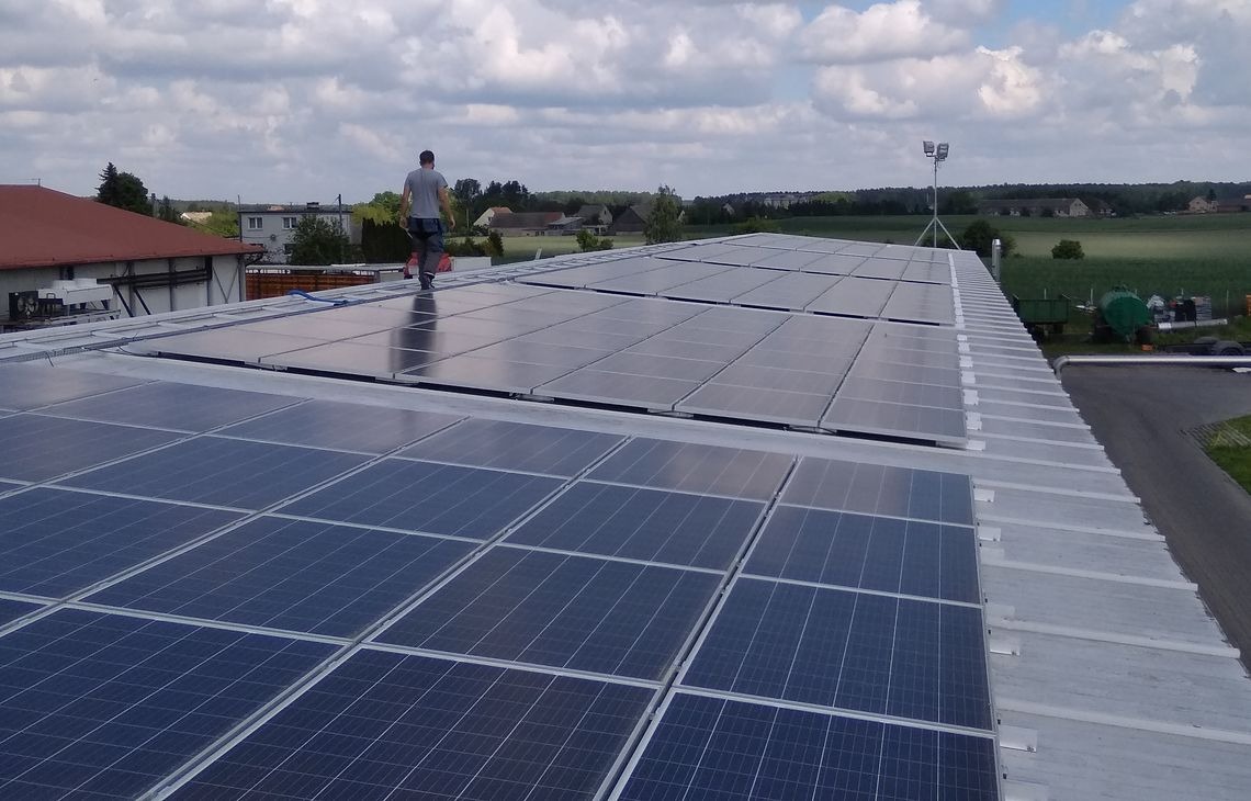 Instalacja paneli fotowoltaicznych na dachu budynku gospodarczego w słoneczny dzień. Widoczny pracownik na panelach i wiejski krajobraz w tle.