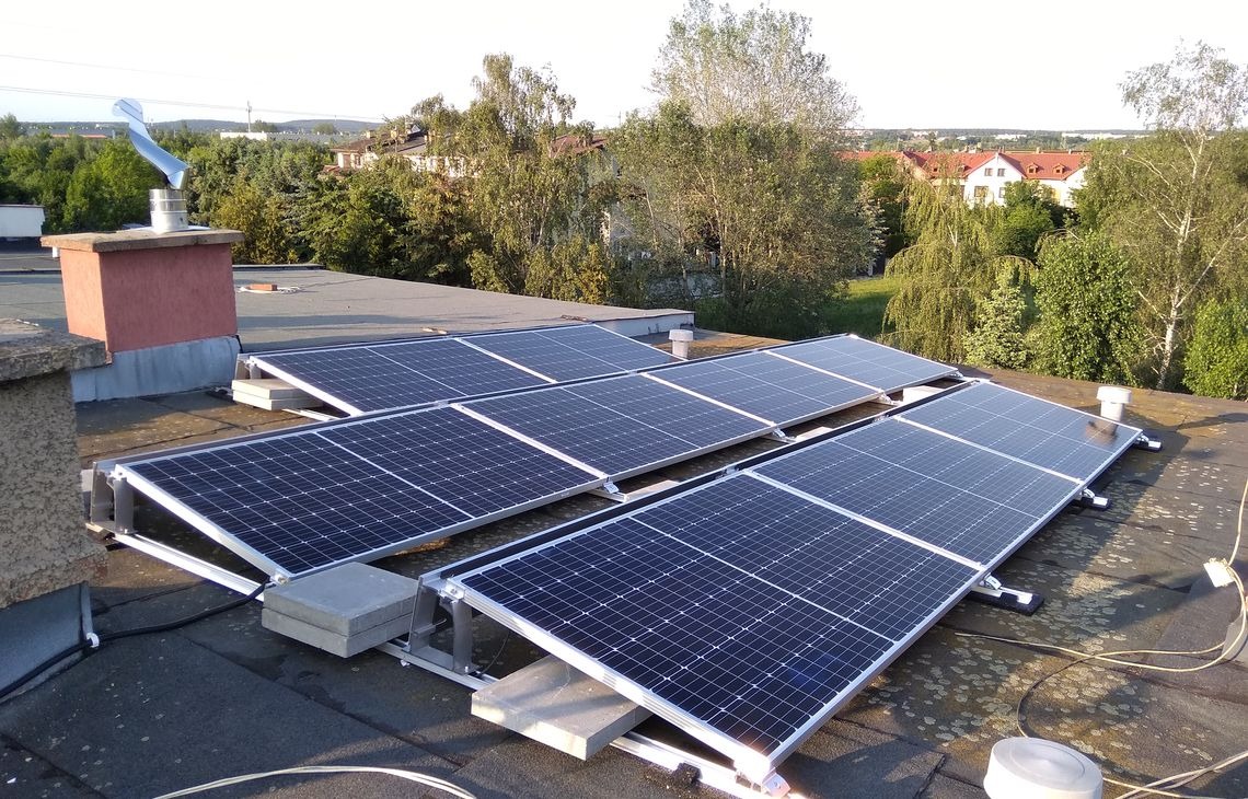Panele fotowoltaiczne na płaskim dachu z widocznym kominem i turbiną wiatrową w tle, w słoneczny dzień. Montaż paneli na specjalnej konstrukcji wsporczej.
