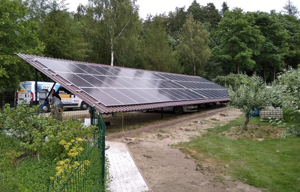Zadaszenie z paneli fotowoltaicznych nad miejscami parkingowymi, częściowo zasłonięte zielenią. W tle zaparkowany biały samochód i bus firmy instalacyjnej.