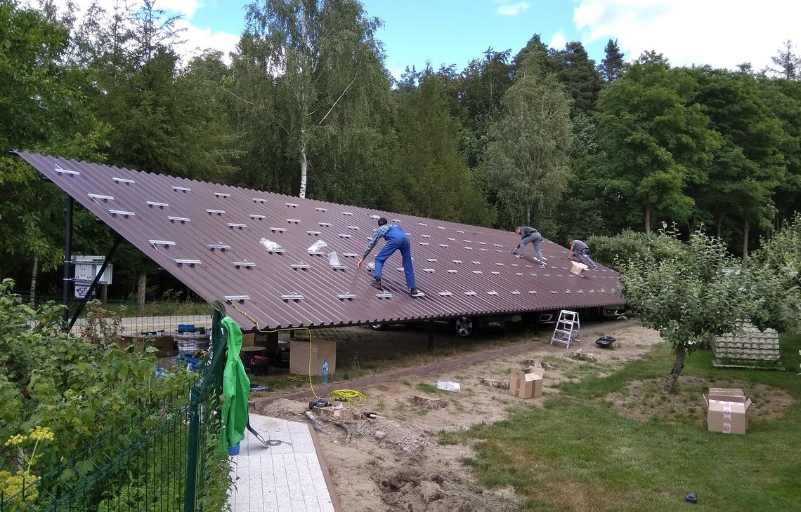 Montaż konstrukcji wsporczej pod panele fotowoltaiczne na gruncie. Trzech monterów w trakcie pracy na brązowym, falistym dachu konstrukcji. Widok z boku, zieleń w tle.