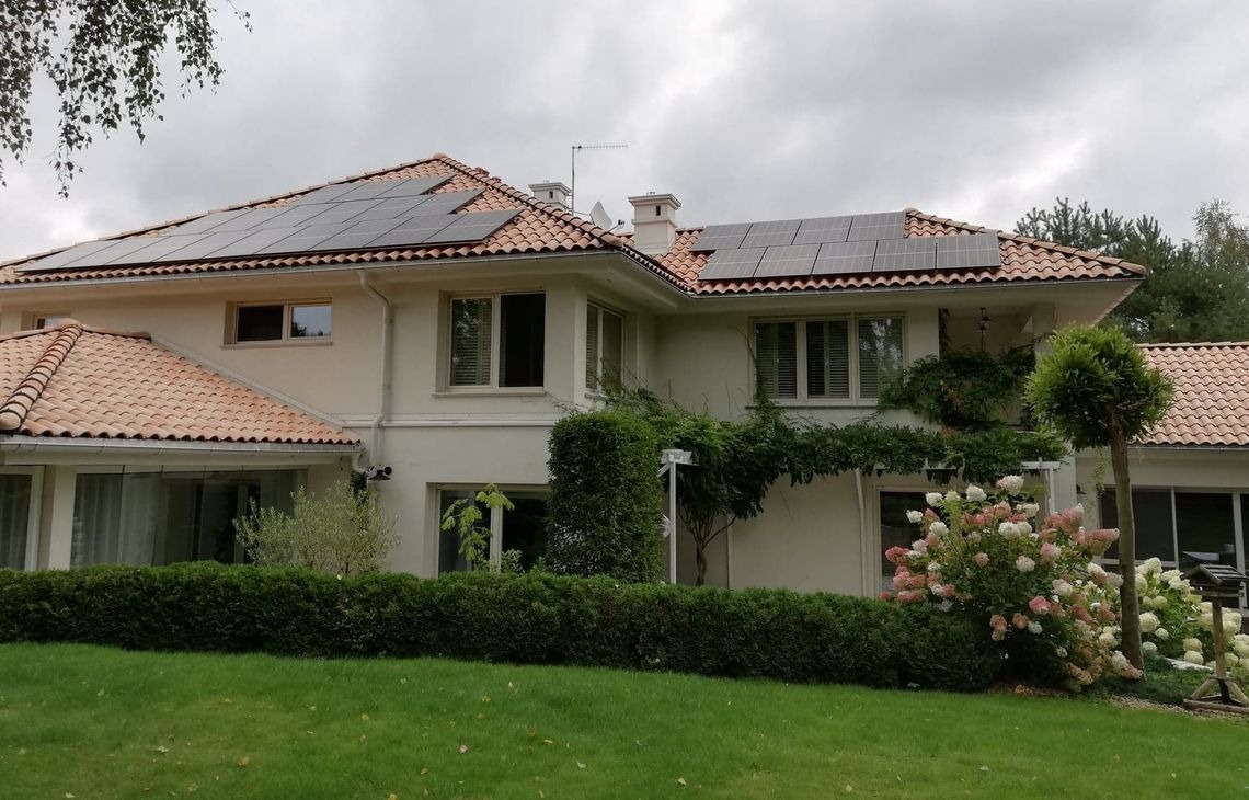 Dom jednorodzinny z panelami fotowoltaicznymi na dachu z dachówką ceramiczną. Widok z poziomu trawnika, pochmurne niebo.