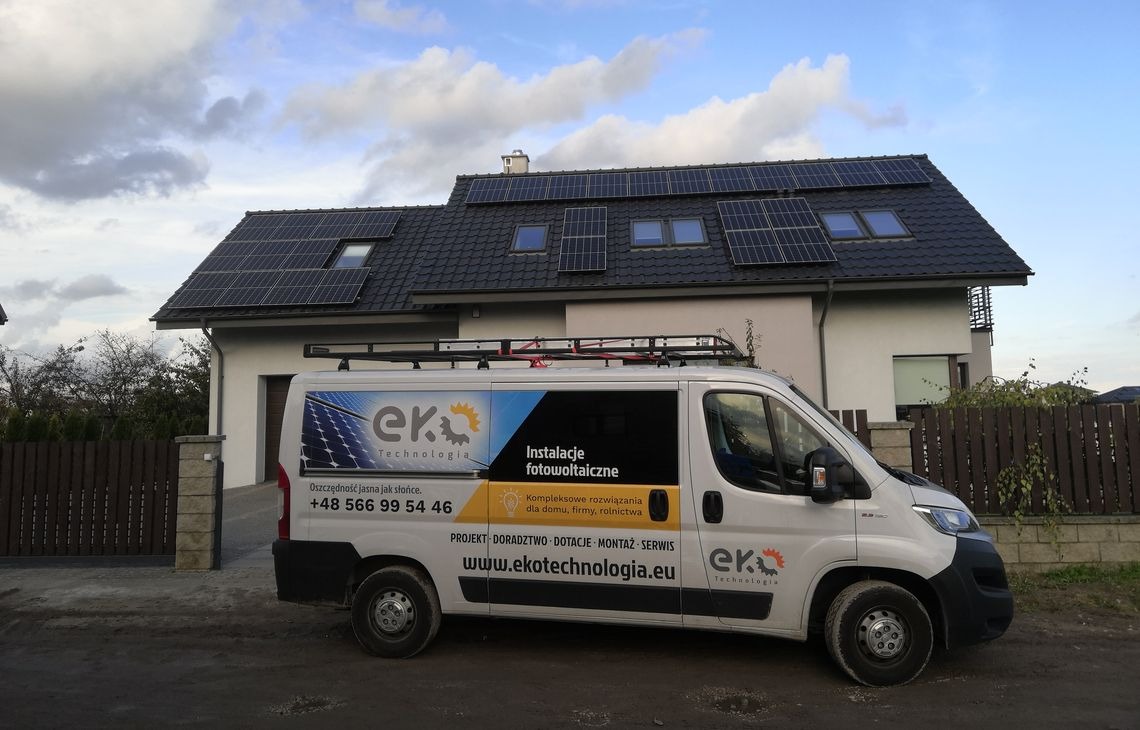 Dom z panelami fotowoltaicznymi na dachu, obok zaparkowany biały bus firmy Eko Technologia z reklamą instalacji fotowoltaicznych. Pochmurne niebo.