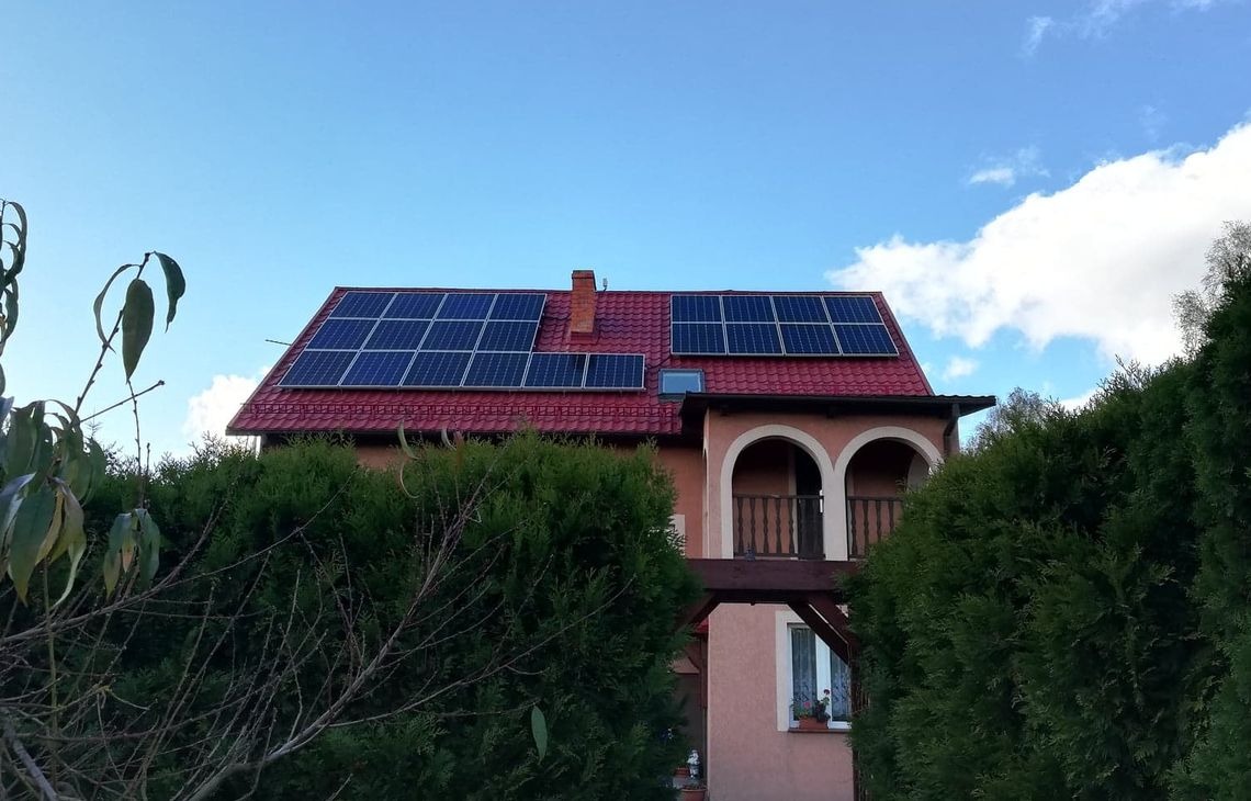 Dom jednorodzinny z panelami fotowoltaicznymi na czerwonym dachu, widoczny zza zielonych krzewów, z balkonem z łukowatymi wejściami i fragmentem błękitnego nieba.
