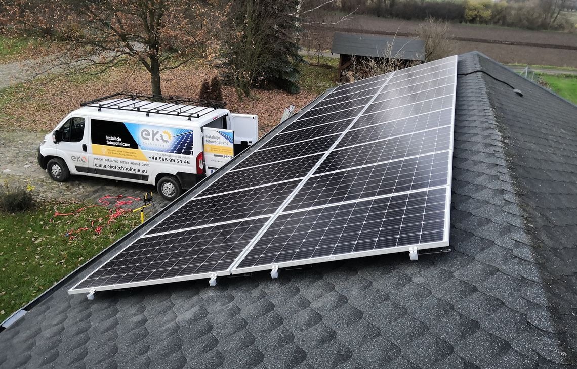 Panel fotowoltaiczny na dachu z bitumicznej dachówki, obok biały bus z logo firmy instalacyjnej. Widok z góry, pochmurny dzień.