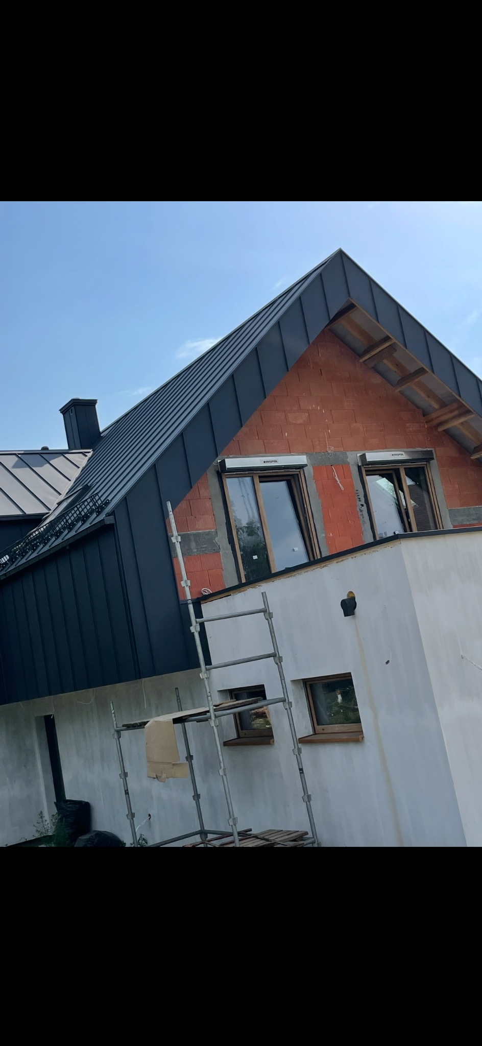 Nowoczesny dom w trakcie budowy z ciemnym dachem z blachy na rąbek stojący i oknami z roletami zewnętrznymi. Widoczne rusztowanie i fragmenty muru.