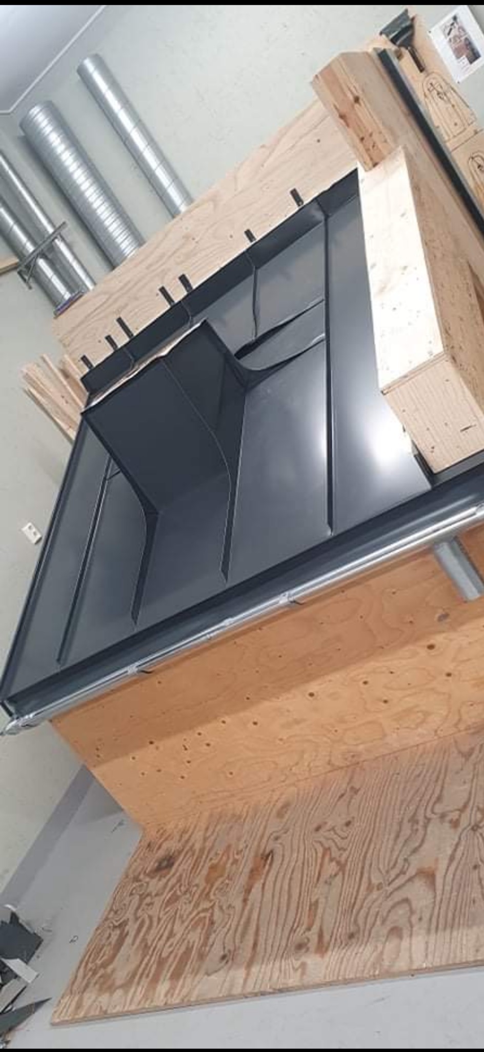 Szara, metalowa konstrukcja rynny dachowej w warsztacie, wsparta na drewnianych elementach. Widoczne rury wentylacyjne w tle. Nietypowy kąt ujęcia.