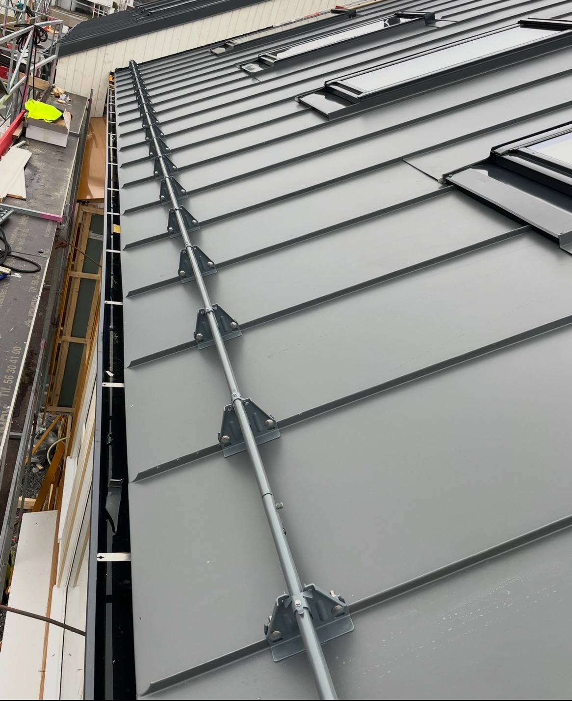 Szary dach z blachy na rąbek stojący, zabezpieczony systemem odśnieżania. Widoczne okna dachowe i fragment rusztowania. Ujęcie z boku, podkreślające linię dachu.