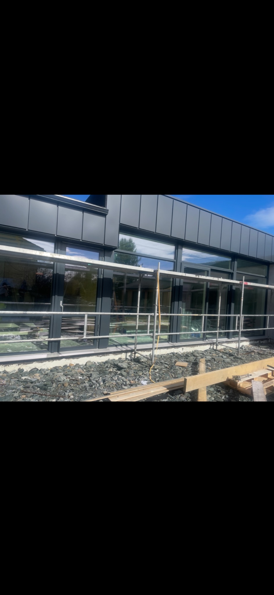 Nowoczesny budynek z szarym dachem i elewacją, widoczny fragment rusztowania i gruz przy ścianie, sugerujący prace wykończeniowe dachu lub elewacji.