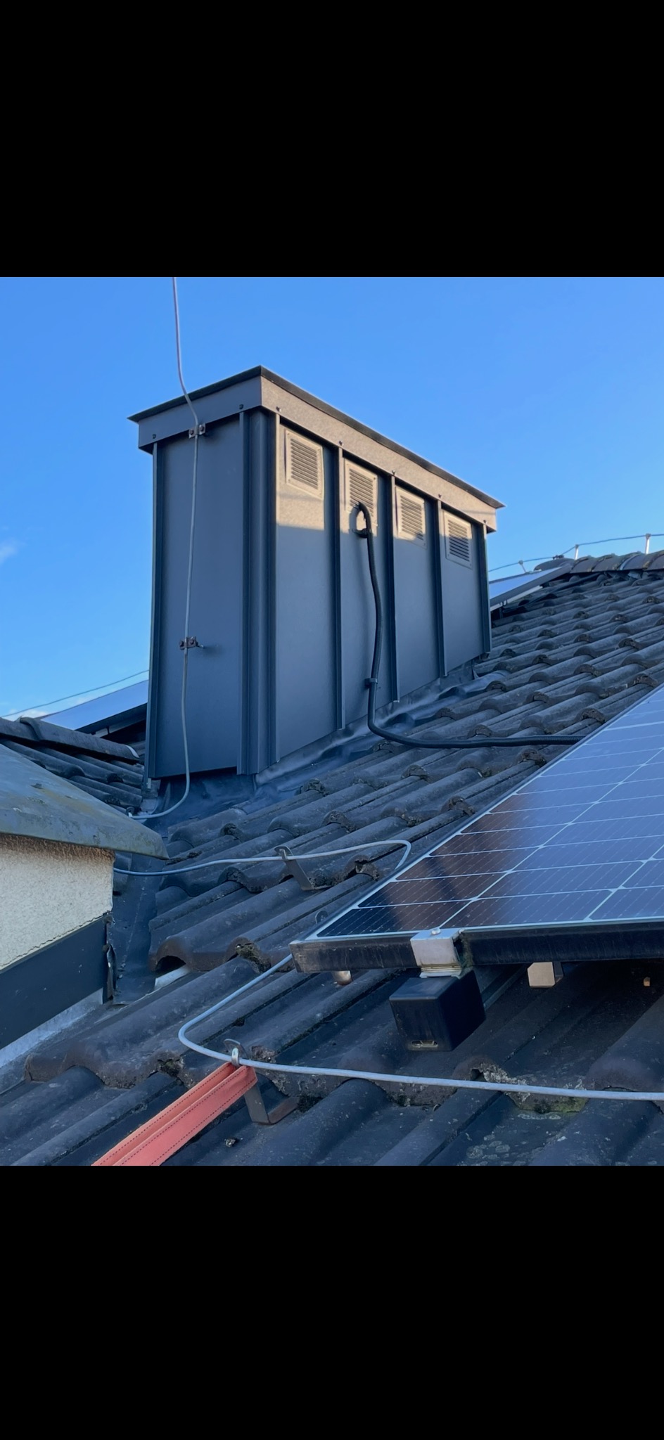 Szary komin wentylacyjny na dachu z dachówki ceramicznej, widoczne panele słoneczne i przewody. Błękitne niebo w tle. Komin z kratkami wentylacyjnymi.