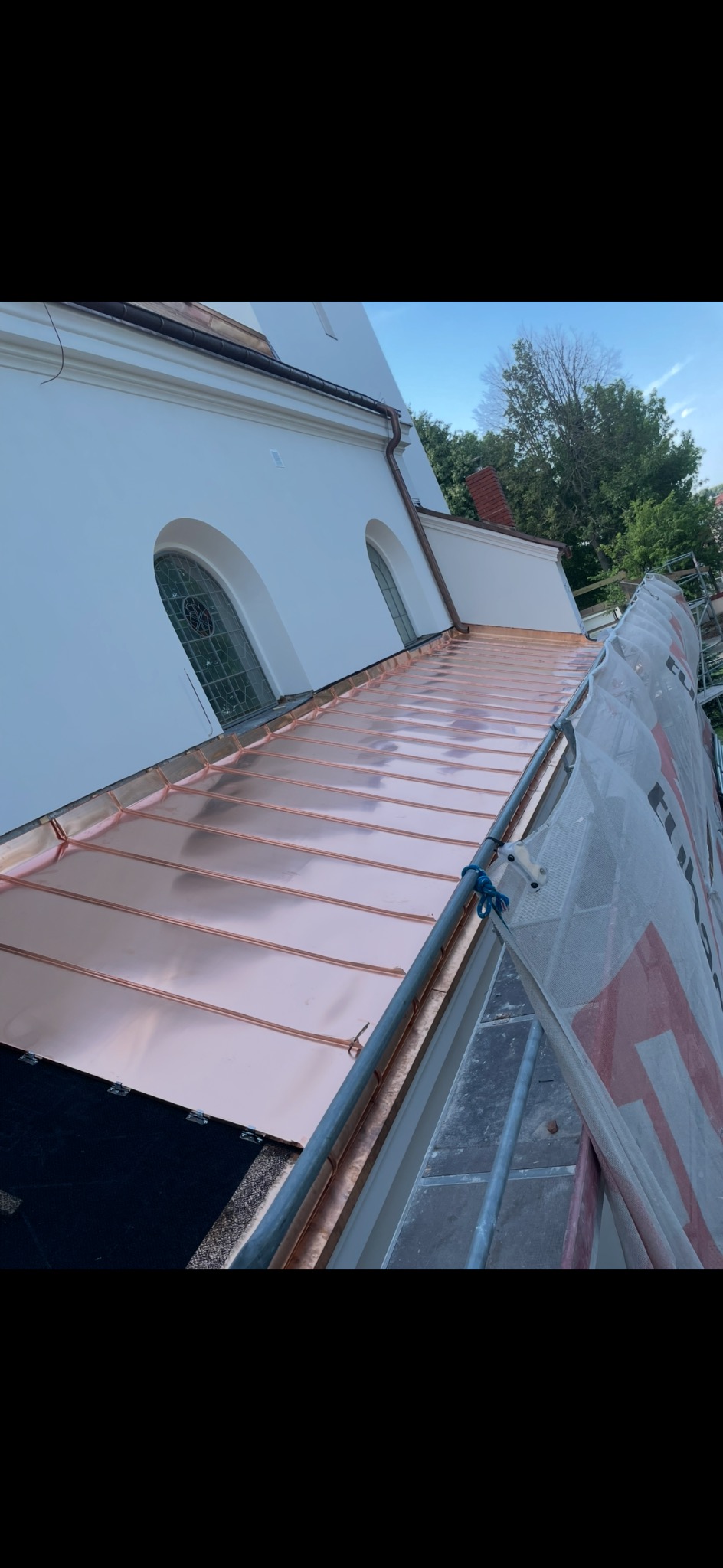 Błyszczący, miedziany dach kościoła w trakcie krycia, widoczny ze rusztowania. Nowe pokrycie dachu z blachy miedzianej, jasne niebo w tle.