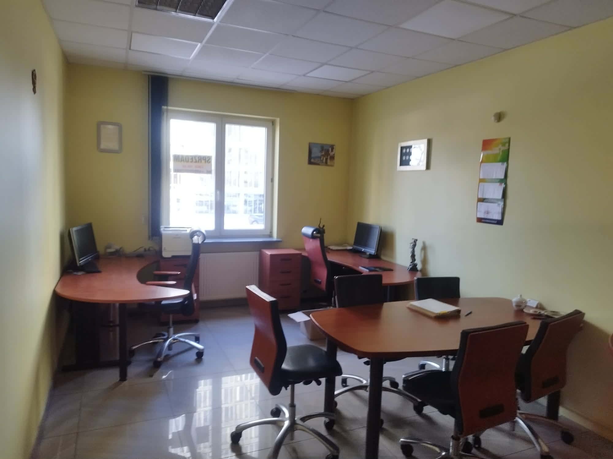 Jasne biuro z żółtymi ścianami, wyposażone w biurka, krzesła obrotowe i komputery. Widoczne okno z zasłoną i elementy dekoracyjne na ścianach. Ustawienie mebli sprzyja pracy zespołowej.