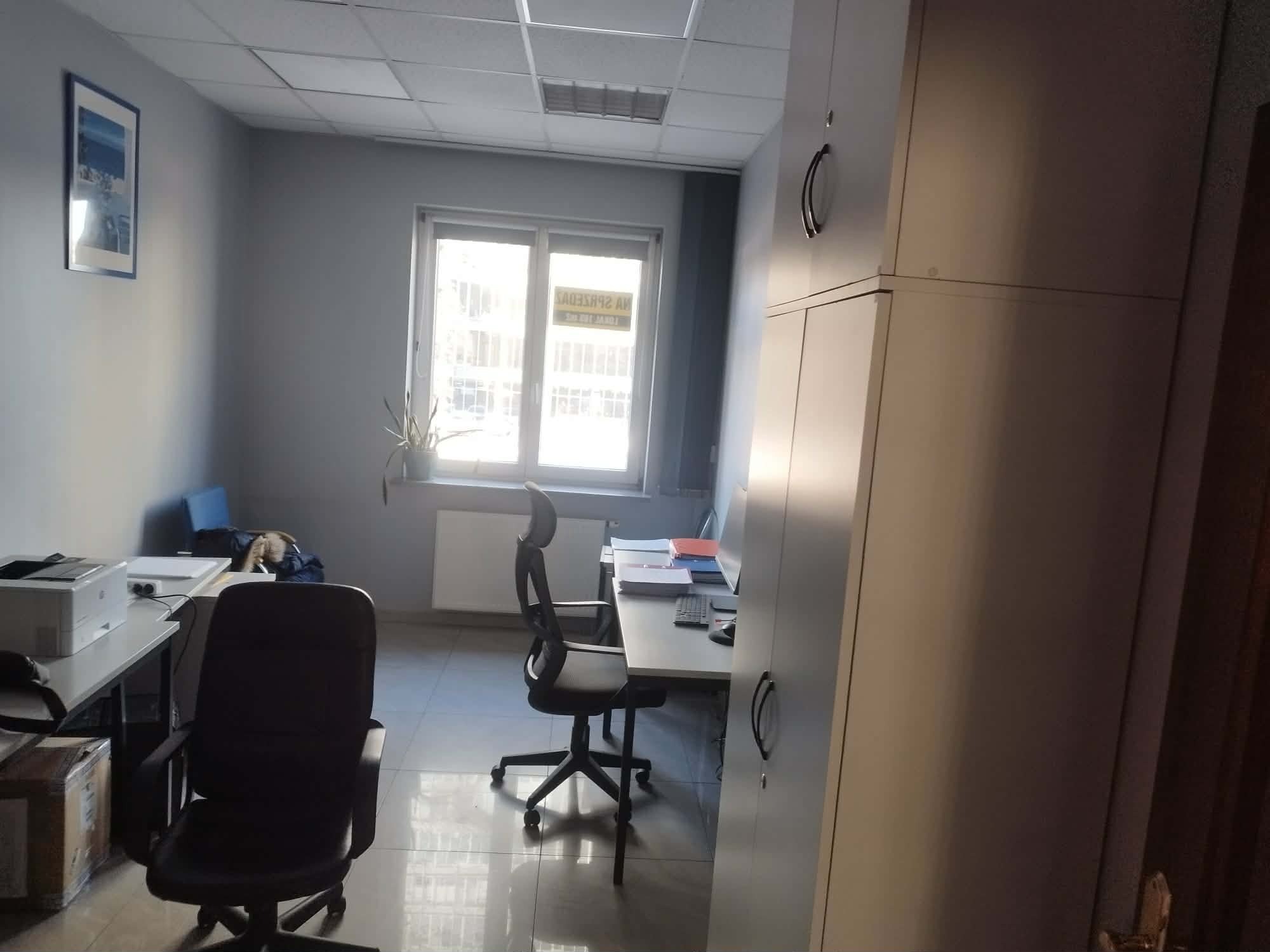 Jasne biuro z biurkami, krzesłami i szafą. Widoczne okno z widokiem na zewnątrz. Minimalistyczny wystrój, szare ściany, płytki na podłodze. W tle obraz na ścianie.