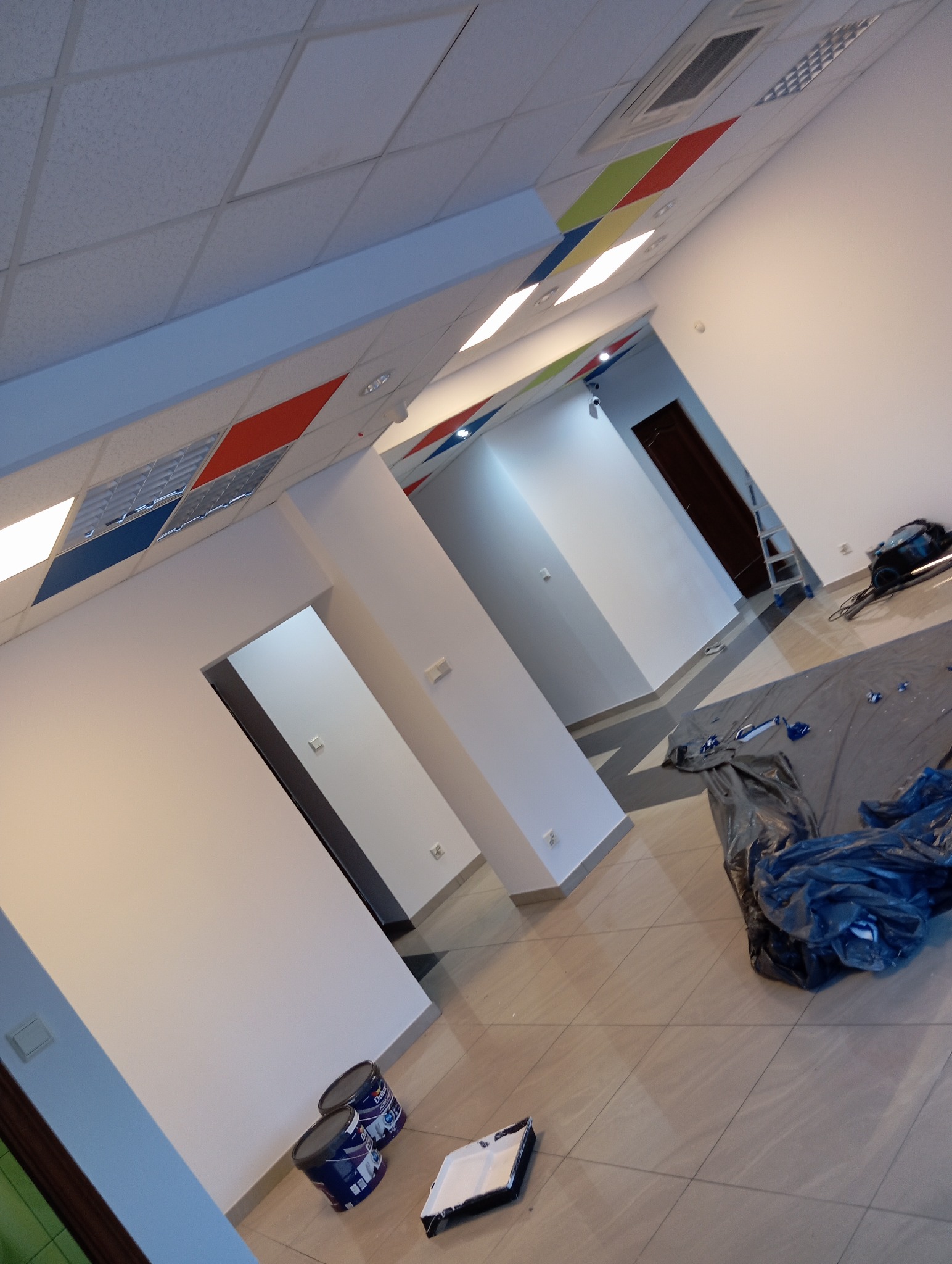 Wnętrze biura w trakcie malowania: białe ściany, kolorowe akcenty na suficie, pędzle i farby na podłodze. Widoczny fragment drabiny i zabezpieczona folią podłoga.