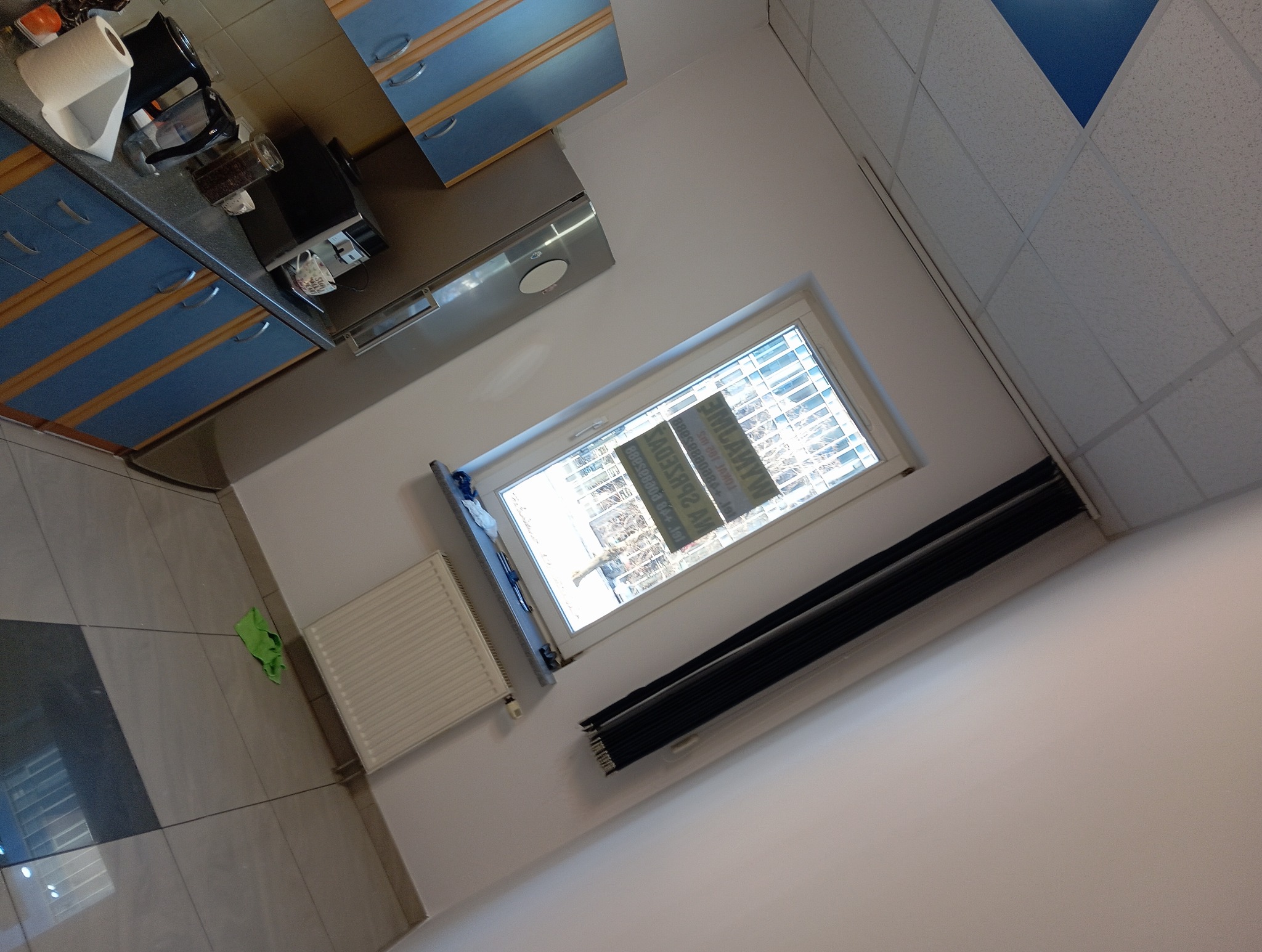 Wnętrze biura z oknem, grzejnikiem i roletami. Widoczny fragment kuchni z niebieskimi szafkami. Ujęcie z perspektywy sufitu, obrócone.