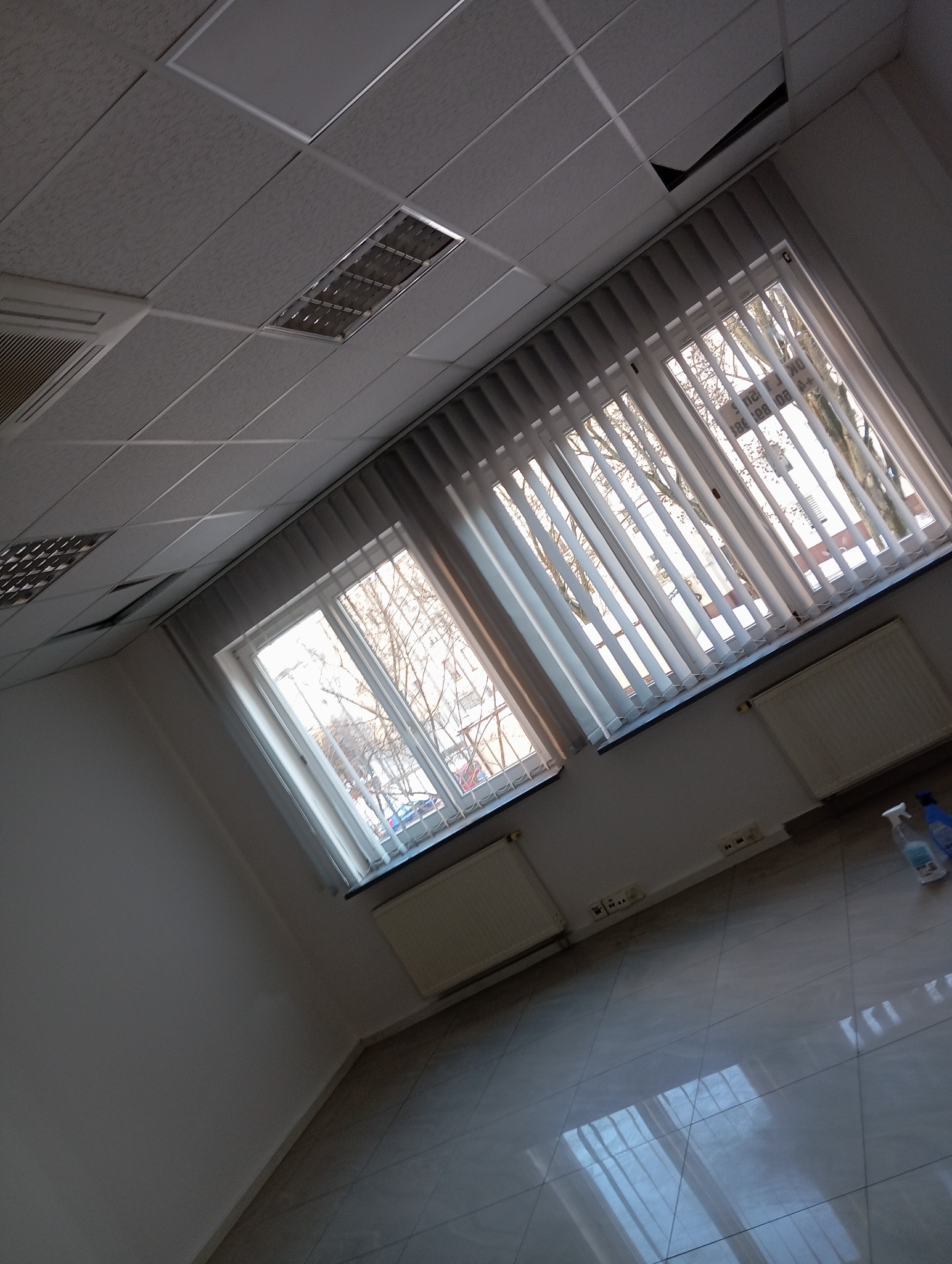 Wnętrze biura z dwoma oknami z żaluzjami, widoczne grzejniki, płytki podłogowe i fragment sufitu podwieszanego. Ukośny kadr, butelki ze środkiem czyszczącym w rogu.