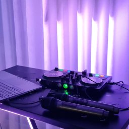 DJ PIORUN - Stanowisko DJ-a z laptopem, mikserem i mikrofonami, oświetlone fioletowym światłem na tle białej zasłony. Widoczny sprzęt audio i komputer.