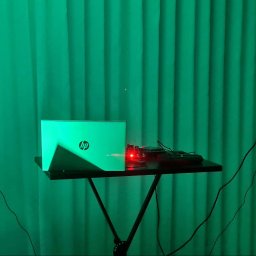 DJ PIORUN - Dwa monitory studyjne Focal na statywach, laptop HP i sprzęt audio na stoliku, oświetlone zielonym światłem na tle zielonej zasłony. Scena gotowa do nagrywania muzyki.