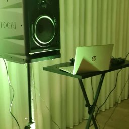 DJ PIORUN - Sprzęt nagłośnieniowy: głośnik Focal na statywie i laptop HP na czarnym stoliku, oświetlone zielonym światłem, na tle białych zasłon. Przygotowanie do występu.