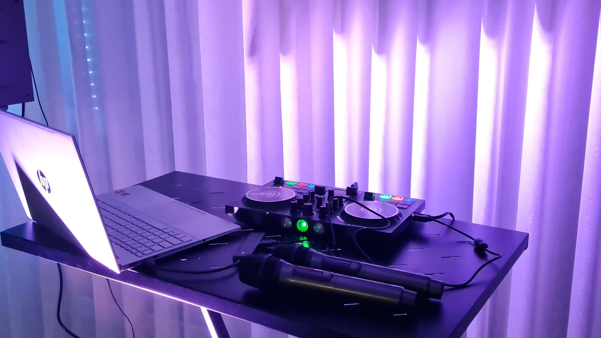 Stanowisko DJ-a z laptopem, mikserem i mikrofonami, oświetlone fioletowym światłem na tle białej zasłony. Widoczny sprzęt audio i komputer.