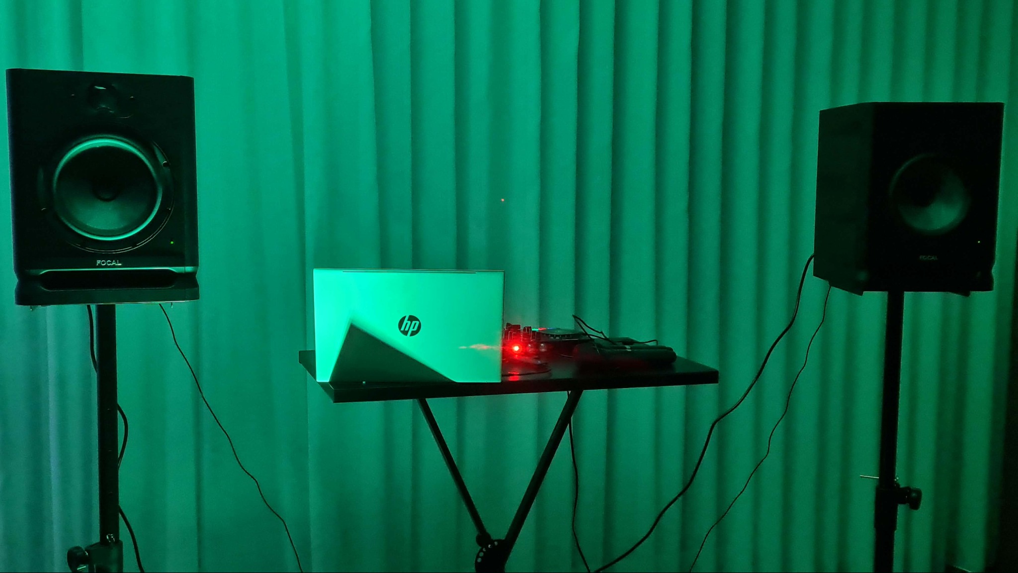 Dwa monitory studyjne Focal na statywach, laptop HP i sprzęt audio na stoliku, oświetlone zielonym światłem na tle zielonej zasłony. Scena gotowa do nagrywania muzyki.