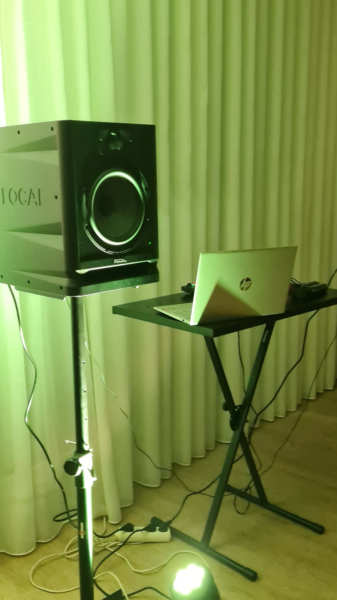 Sprzęt nagłośnieniowy: głośnik Focal na statywie i laptop HP na czarnym stoliku, oświetlone zielonym światłem, na tle białych zasłon. Przygotowanie do występu.