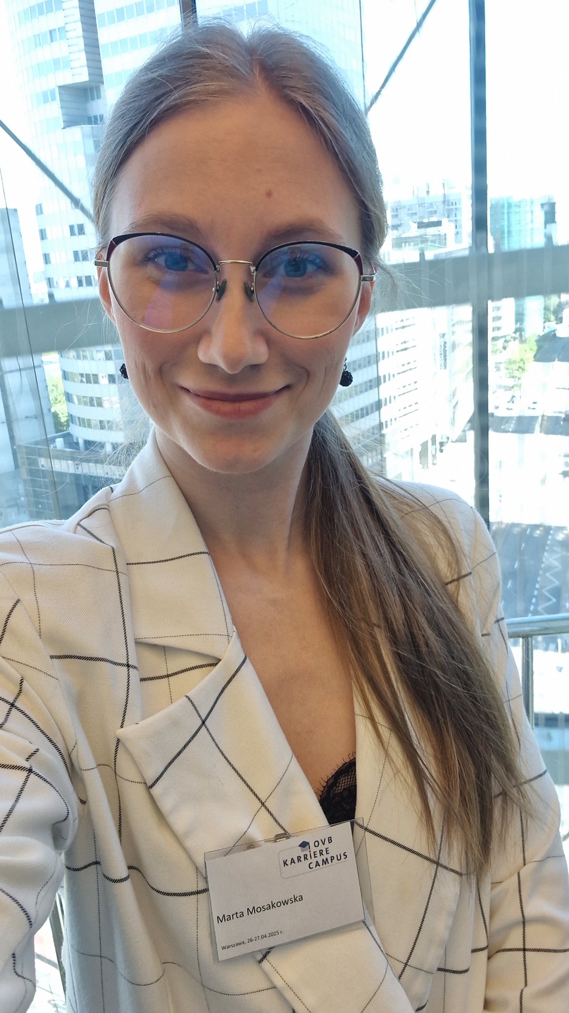 Portret uśmiechniętej kobiety w okularach i marynarce w kratę, z identyfikatorem OVB Karriere Campus na tle nowoczesnego biurowca.