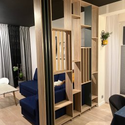 DAWID PAWLAK - Drewniany regał działowy z półkami w salonie, oddzielający strefę wypoczynkową z niebieską sofą. Elementy w kolorze drewna i grafitu, nowoczesny design.
