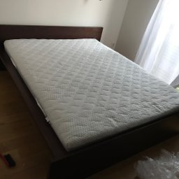 DAWID PAWLAK - Ciemnobrązowe łóżko z białym materacem w pokoju z drewnianą podłogą. W tle okno z białymi zasłonami i fragment grzejnika. Na podłodze szczotka i folia.