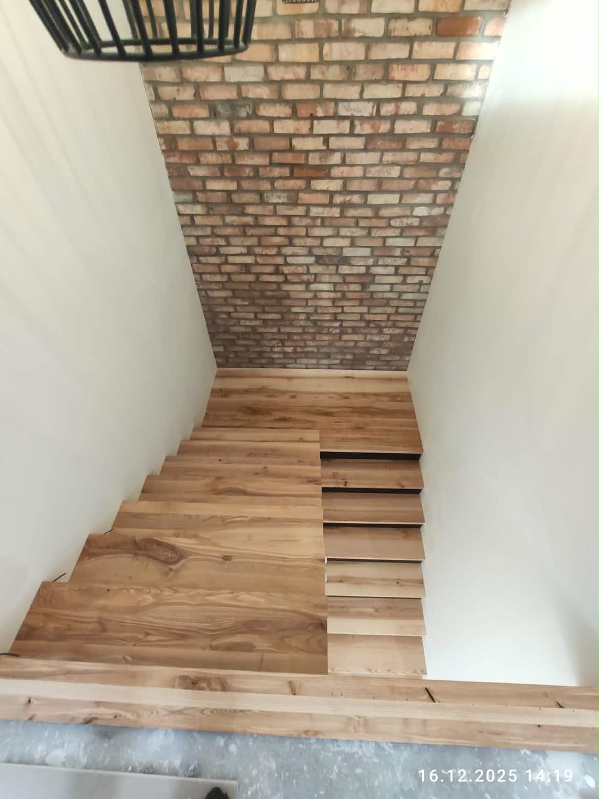 Drewniane schody z widoczną strukturą drewna, prowadzące w dół, z ceglaną ścianą w tle i nowoczesną lampą. Ujęcie z góry.