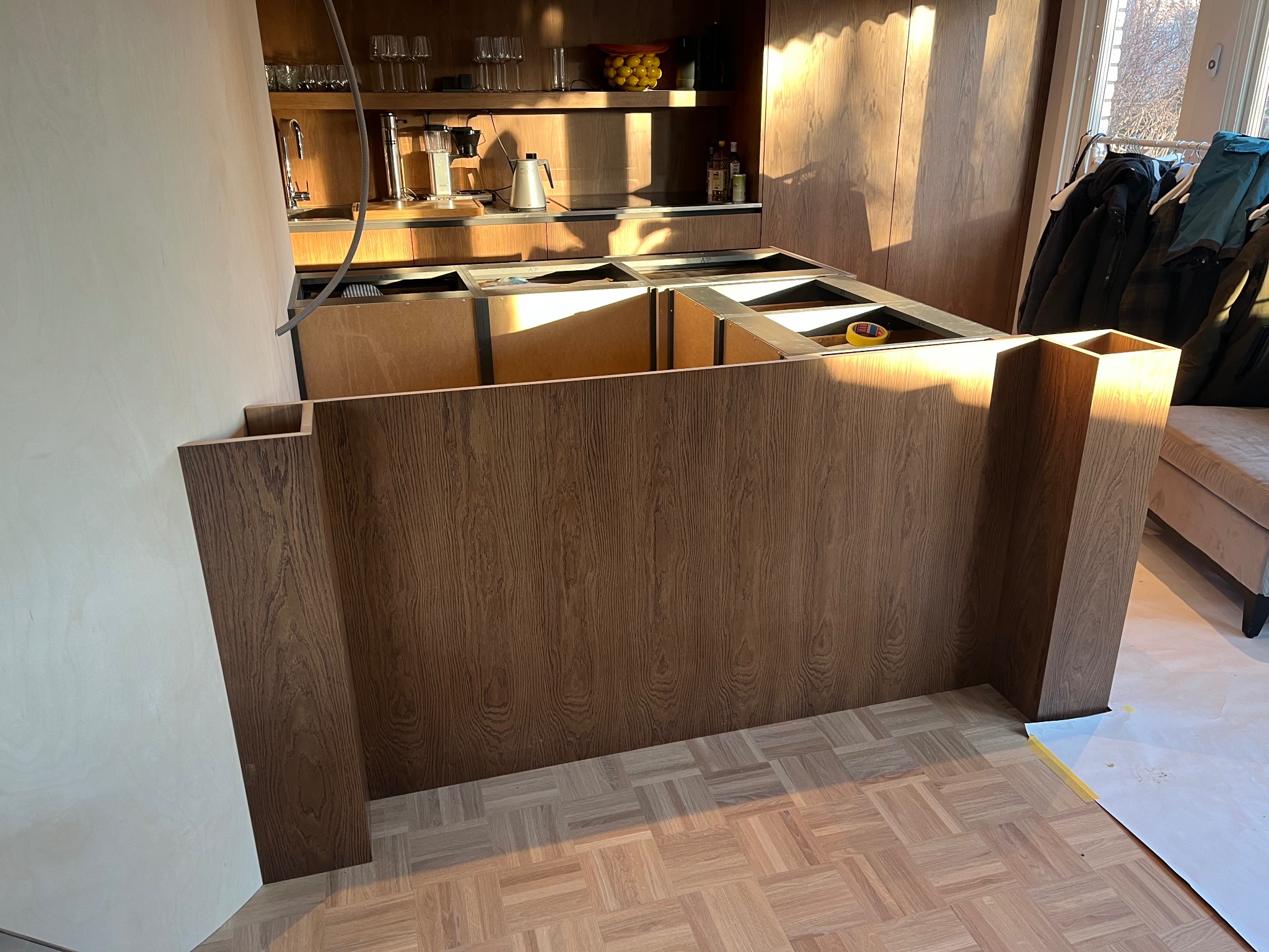 Montaż drewnianej wyspy kuchennej z widoczną strukturą drewna i parkietem w jodełkę. W tle elementy wyposażenia kuchni i wpadające promienie słoneczne.