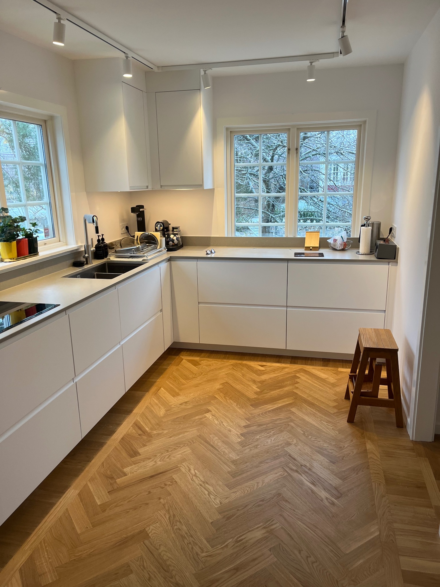 Jasna kuchnia z białymi frontami i drewnianą podłogą ułożoną w jodełkę. Widoczne okna z widokiem na drzewa i oświetlenie punktowe na suficie. Minimalistyczny design.