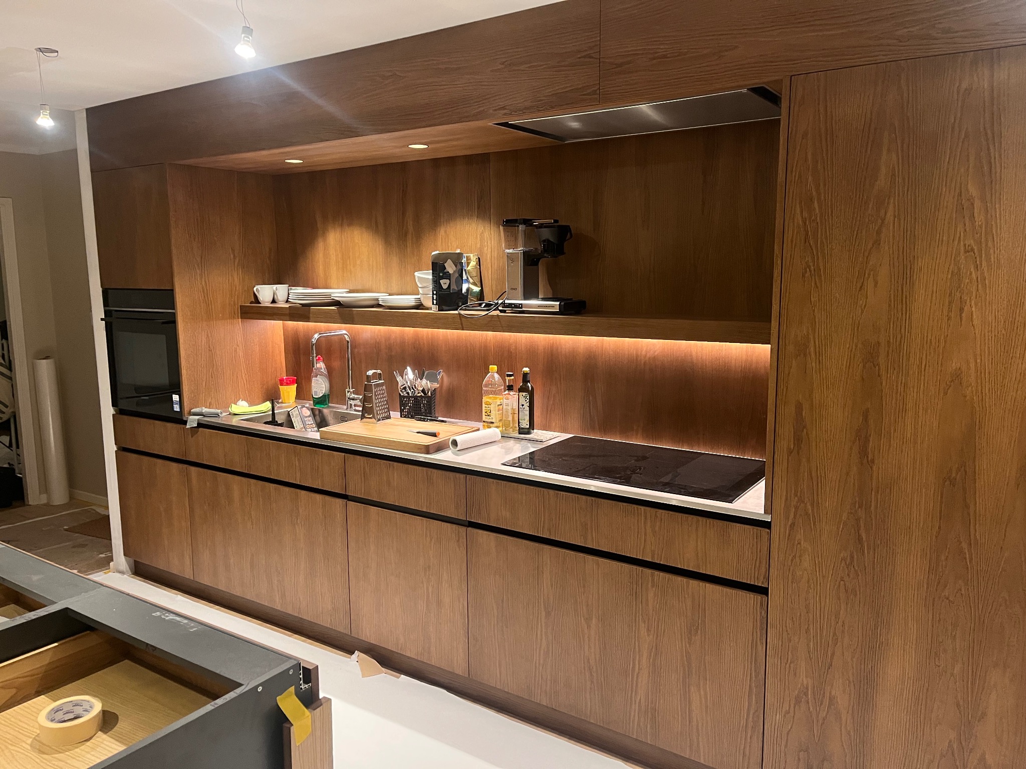 Nowoczesna kuchnia z drewnianymi frontami, oświetleniem LED pod szafkami i sprzętem AGD na półce. Częściowo widoczny montaż dolnych szafek.