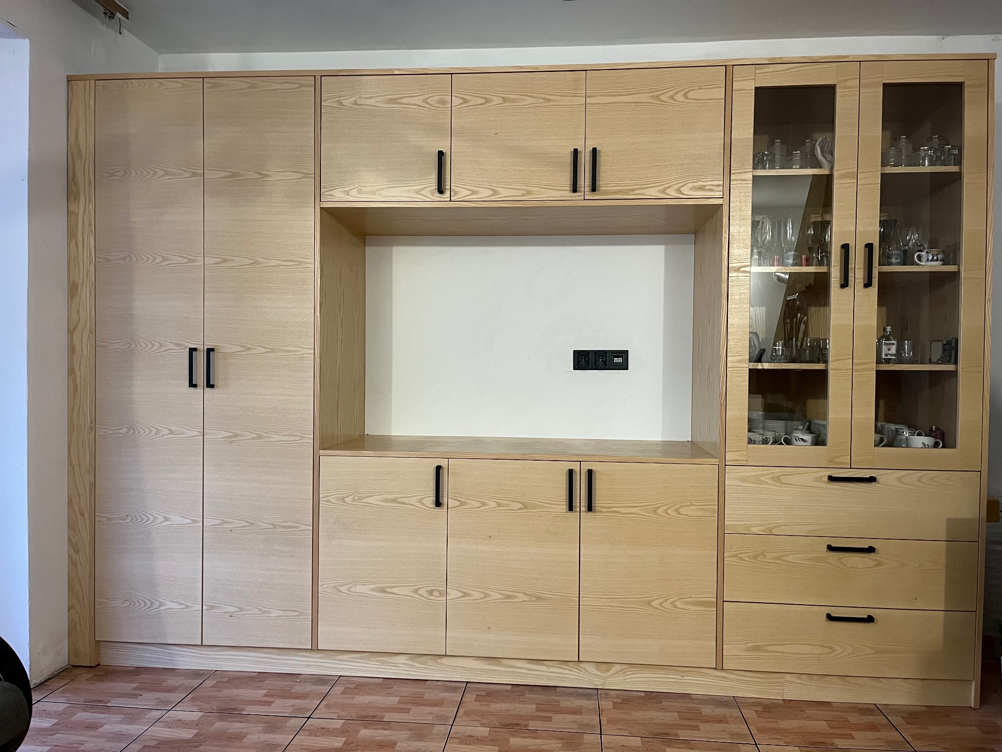 Jasna, drewniana zabudowa meblowa z szafkami, szufladami i przeszkloną witryną, z czarnymi, minimalistycznymi uchwytami, na tle kafelkowanej podłogi.