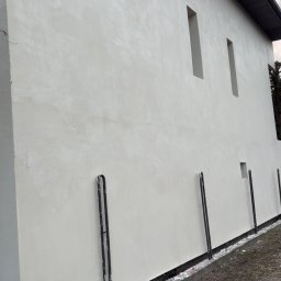 Krzysztof Gawarski - Jasna, gładka elewacja budynku z wąskimi, pionowymi oknami. Widoczne metalowe wsporniki przy podstawie ściany i fragment korony drzewa w tle.