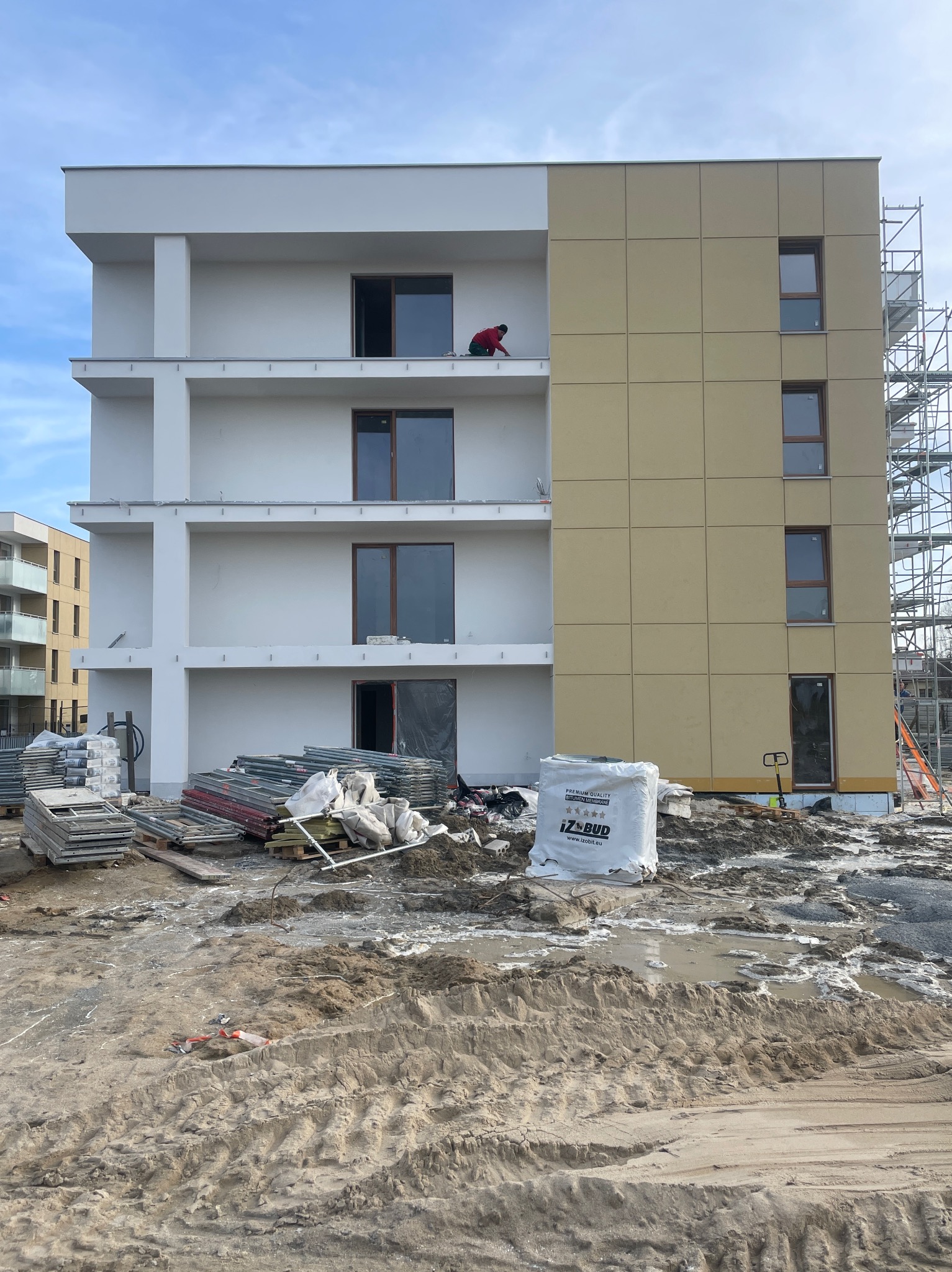 Wielopiętrowy budynek w trakcie prac elewacyjnych, z widocznymi rusztowaniami, materiałami budowlanymi i pracownikiem na balkonie. Część elewacji pokryta panelami.