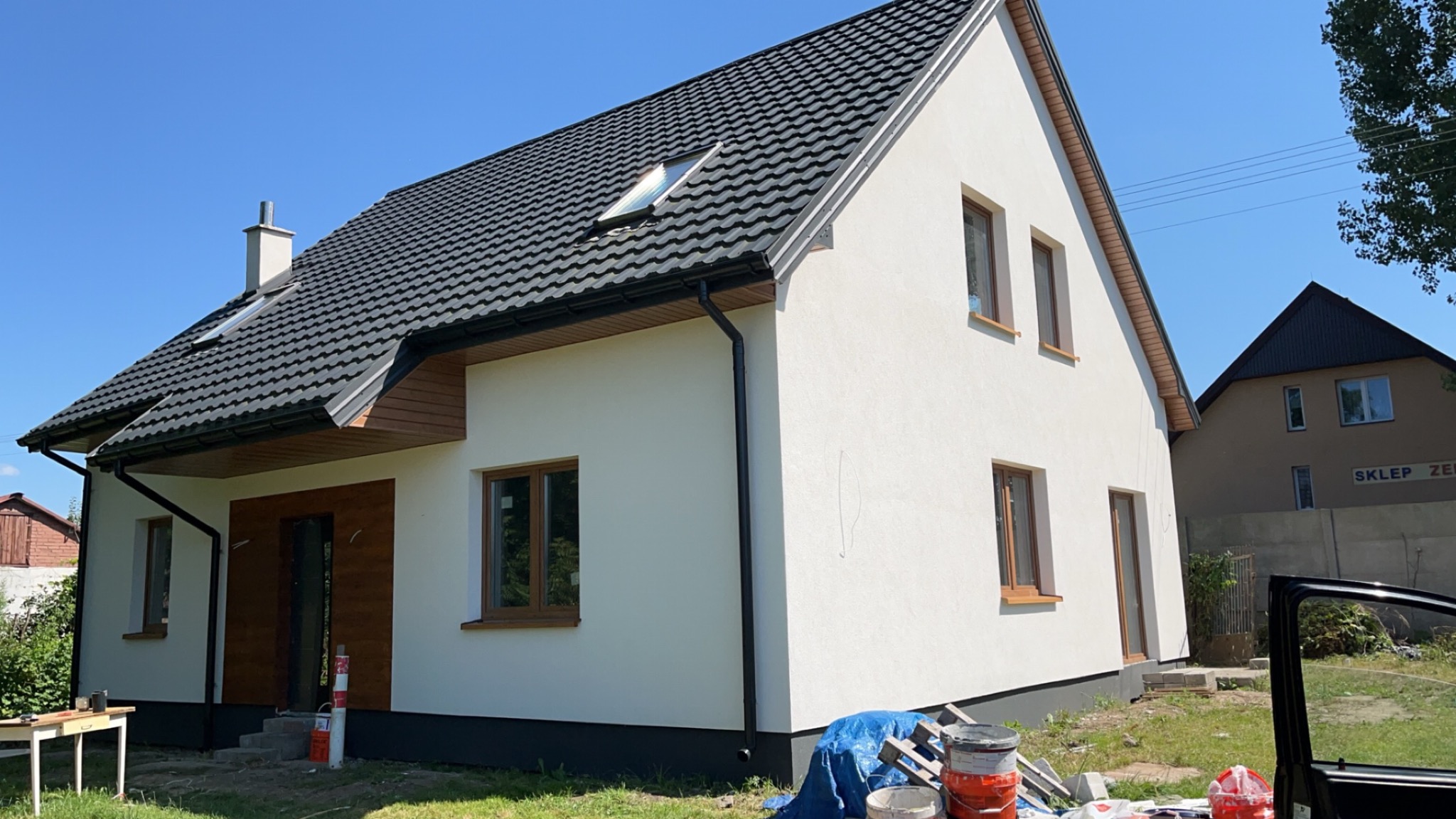 Biała elewacja domu jednorodzinnego z ciemną dachówką, widok z boku, z elementami wykończenia drewnianego i oknami w brązowych ramach. Na pierwszym planie wiaderka z farbą.