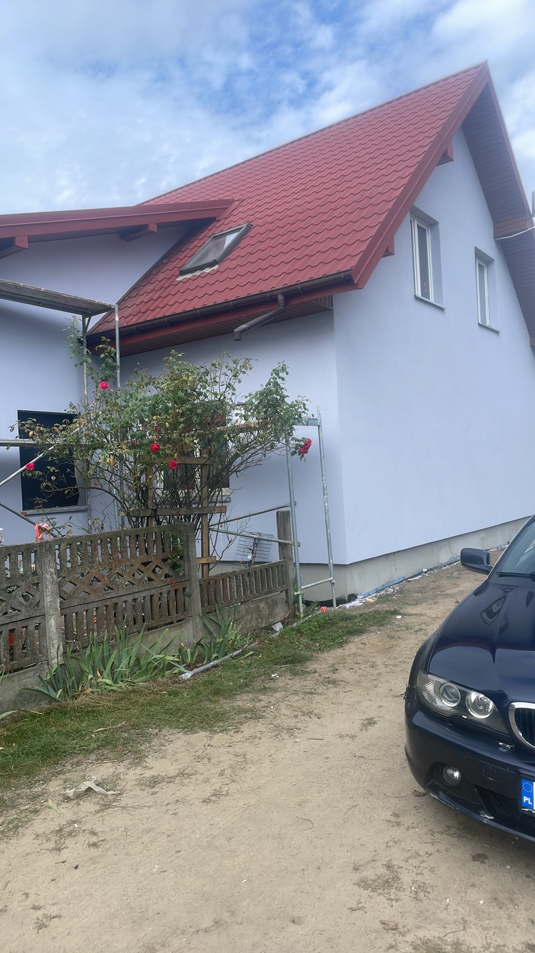 Dom po malowaniu elewacji na jasny kolor, z widocznymi rusztowaniami i fragmentem samochodu. Czerwony dach i świeżo pomalowane ściany.