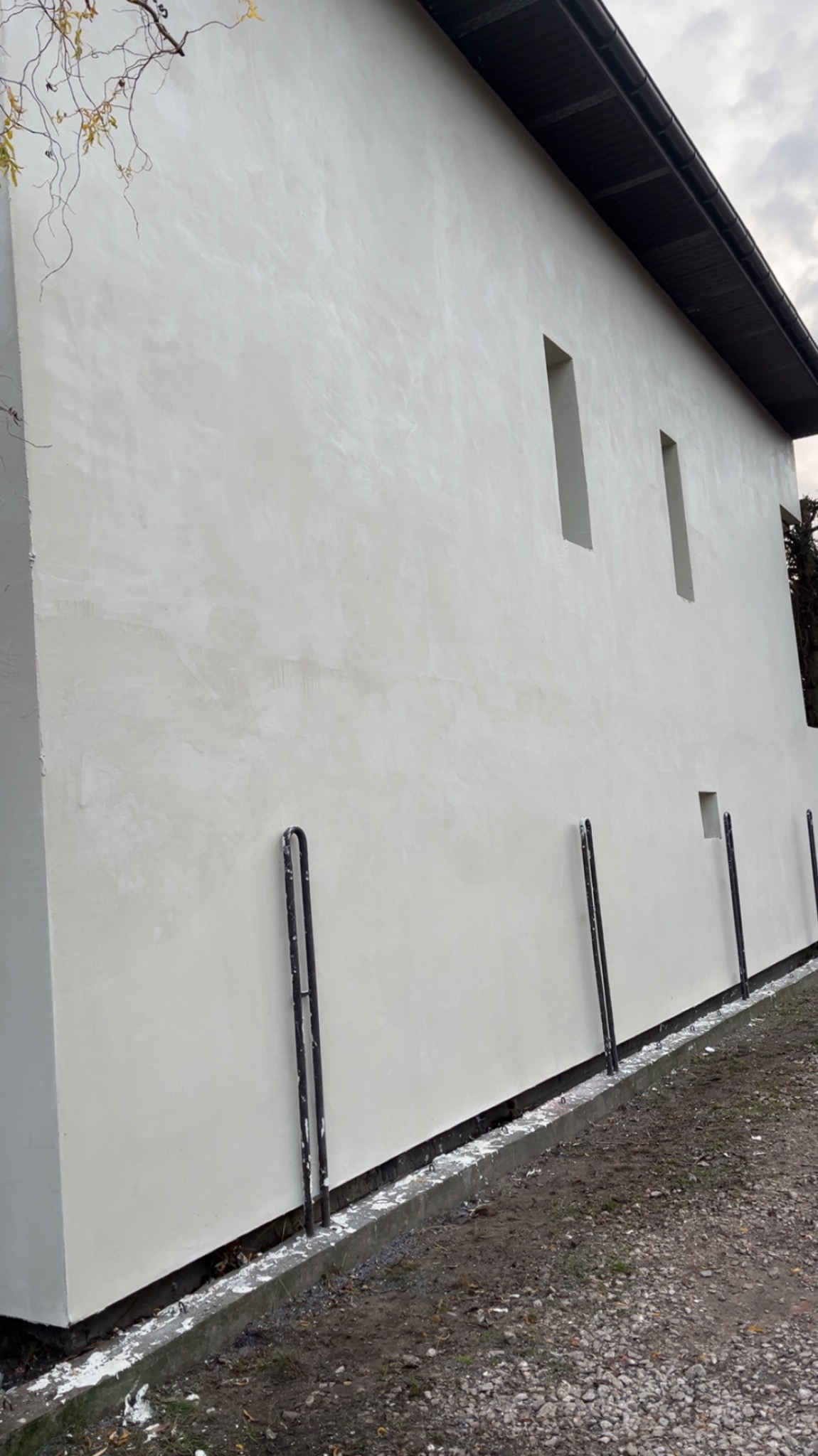 Jasna, gładka elewacja budynku z wąskimi, pionowymi oknami. Widoczne metalowe wsporniki przy podstawie ściany i fragment korony drzewa w tle.