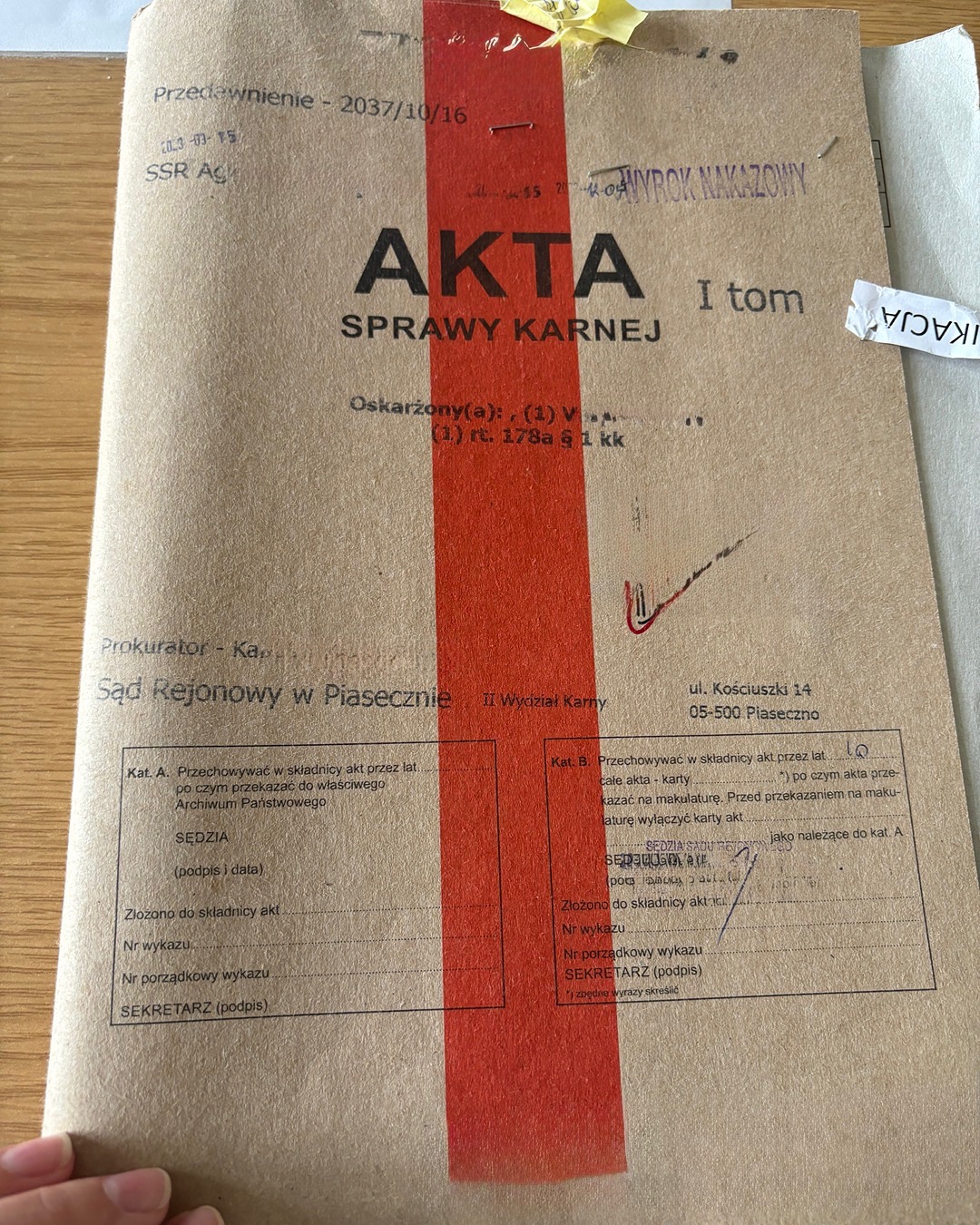 Brązowa teczka akt sprawy karnej z czerwonym paskiem, pieczątkami i odręcznymi notatkami, leżąca na drewnianym blacie. Widoczny fragment dłoni.