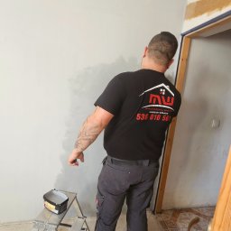 Wykończenia Wnętrz Marcin Wójcik - Mężczyzna w czarnej koszulce z logo firmy maluje szarą farbą ścianę wewnątrz pomieszczenia. Widoczna drabina i puszka z farbą. Częściowo wykończone wnętrze.